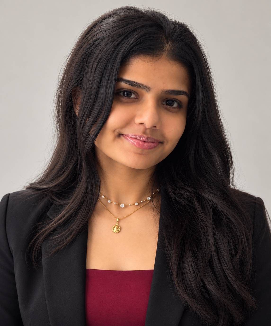 Studio portrait of Keya Patel in a black blazer over a burgundy top, wearing gold necklaces, against a soft gray backdrop